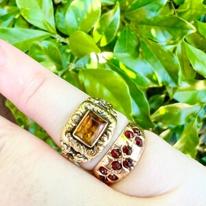 May include: Two gold rings set with gemstones. The top ring features a square, amber-coloured stone in an intricate, ornate design. The lower ring has a band of small, red gemstones.