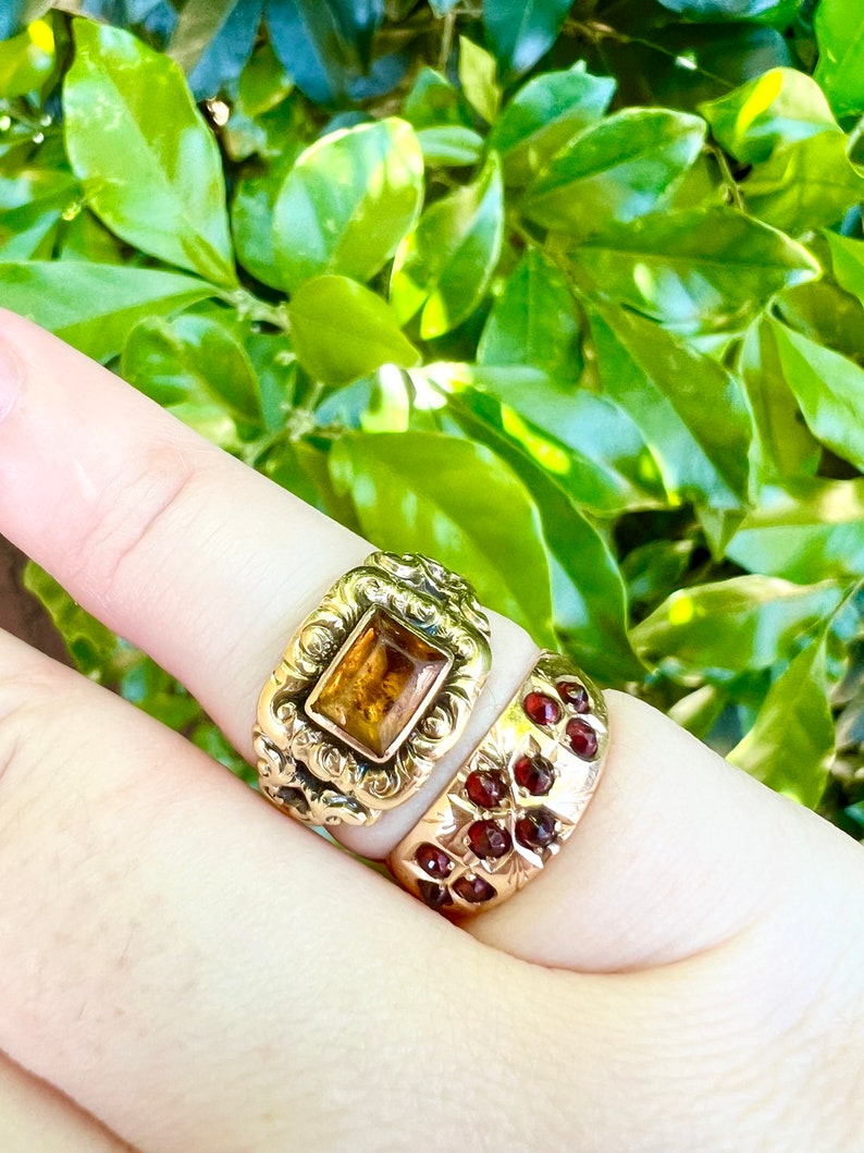 May include: Two gold rings set with gemstones. The top ring features a square, amber-coloured stone in an intricate, ornate design. The lower ring has a band of small, red gemstones.