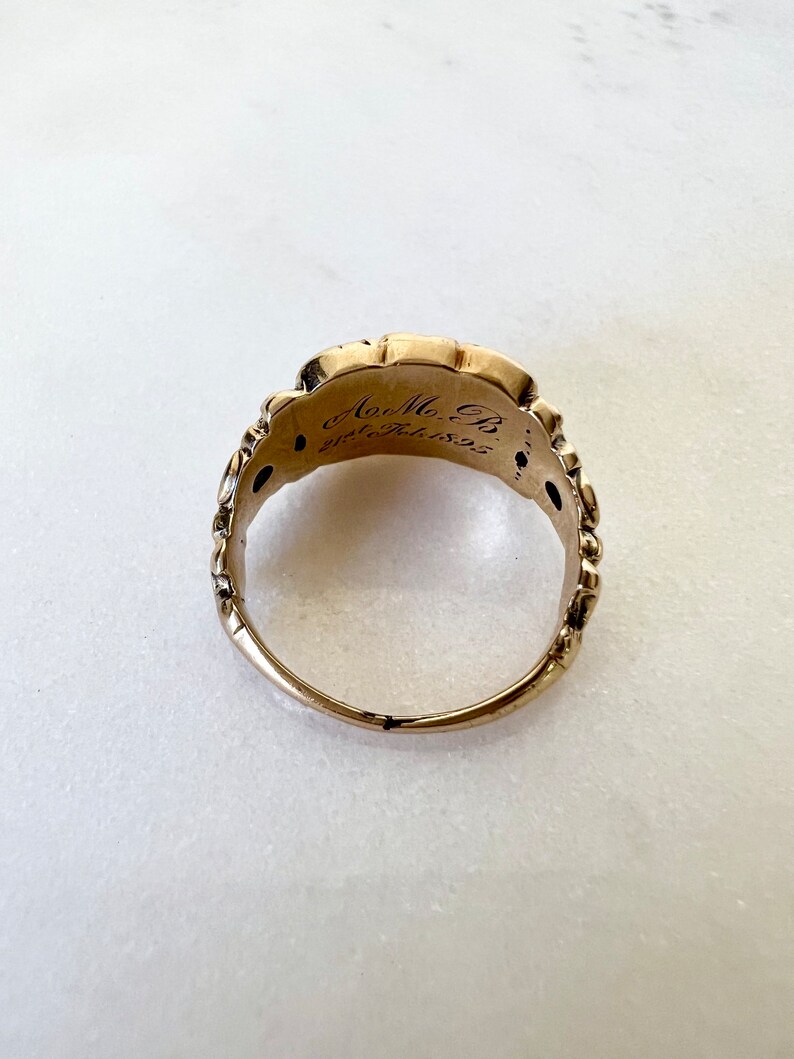 May include: A gold ring with an intricate design and inscription. The inscription reads "A.M.B. 28th Feb 1855".