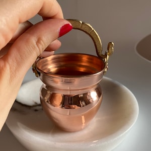 Puede incluir: Una pequeña olla de cobre con un asa de latón. La olla está sobre una superficie de mármol blanco.