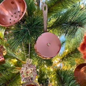 May include: A Christmas tree decorated with copper-coloured ornaments, including a small copper pan and a copper strainer. There is also a clear glass ornament shaped like a gift box.