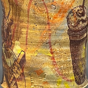 May include: A ceramic mug with a yellow, orange, and brown glaze. The mug has a design of a milkshake with a straw and a person with a hat and a cane.