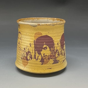 May include: A handmade ceramic mug with a tan base and a textured surface. The mug features a repeating design of purple portraits and tan figures. The rim is a lighter tan color.