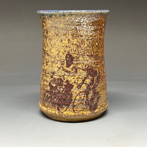 May include: A ceramic vase with a brown and yellow glaze and a blue rim. The vase has a design of people riding bicycles in brown.