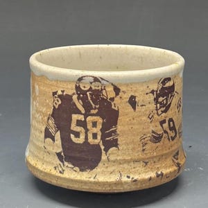 May include: A speckled beige ceramic cup featuring dark brown screen-printed graphics of two football players in uniform. One player wears the number 58 and the other wears the number 59. The cup has a cylindrical shape with a slightly flared rim.
