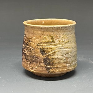 May include: A small, brown ceramic cup with a textured surface. The cup has a wide base and a slightly flared rim.