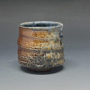 May include: Handmade ceramic tea bowl with a textured surface. The bowl features a brown, blue, and tan color scheme with a unique, organic shape. The exterior has a rough, uneven texture, and a small emblem is visible near the base. A unique piece for tea lovers.