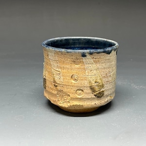 May include: A small, ceramic cup with a light brown glaze and blue rim. The cup has a unique, hand-painted design with white and brown accents.