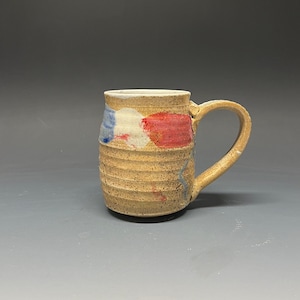 May include: A handmade ceramic mug with a brown glaze and blue, red, and white painted accents. The mug has a rounded body and a handle.