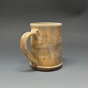 May include: A cylindrical ceramic mug with a speckled tan and brown glaze. The mug features a thick, curved handle attached to the side, a slightly flared rim, and a visible horizontal texture along the body with subtle dark brown markings.