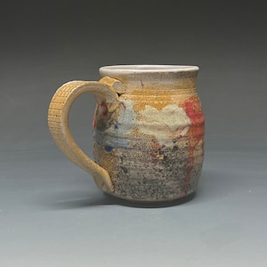 May include: A handmade ceramic mug with a brown, white, and red speckled glaze. The mug has a unique, asymmetrical handle.