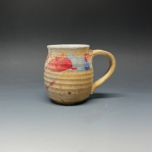 May include: A handmade ceramic mug with a light brown glaze and red and blue paint splatters. The mug has a rounded body and a handle.