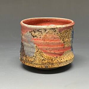 May include: A small, ceramic teacup with a brown, red, and blue glaze. The cup has a textured surface and a slightly flared rim.