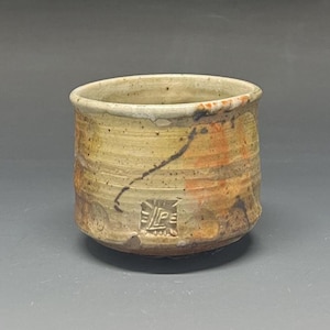 May include: A handmade ceramic cup with a cylindrical shape. The cup features a speckled beige and orange glaze with a dark brown line and a stamped design near the base. The cup is approximately 3 inches tall and 4 inches wide.
