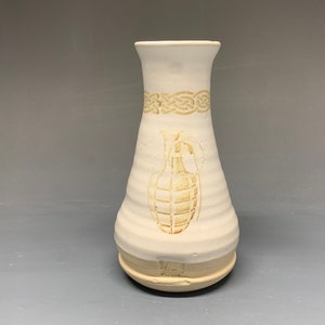 May include: White ceramic vase with a brown design of a grenade and a Celtic knot. The vase has a rounded bottom and a narrow neck.