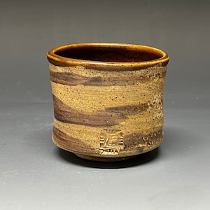 May include: A brown and tan ceramic cup with a striped pattern. The cup has a gold-colored mark on the bottom.