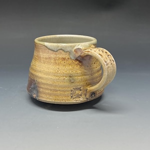 May include: Handmade ceramic mug with a textured, earthy design. The mug features a wide base, a rounded body, and a sturdy handle. The exterior is primarily tan with darker brown accents and a light-colored rim. A small stamp is visible near the base.