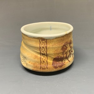 May include: A small, ceramic cup with a light brown glaze and a brown design. The design features a Celtic knot and a stylized animal figure.