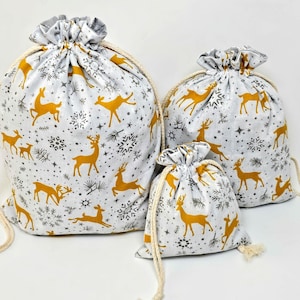May include: Set of three white fabric gift bags with a gold reindeer and snowflake pattern. The bags are different sizes and have a drawstring closure.
