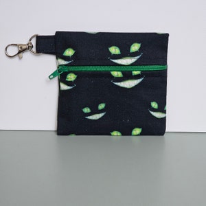 May include: A small, black zippered pouch featuring a repeating pattern of green smiling faces. The zip is green, and a silver metal clasp is attached. The pouch is likely made of fabric.