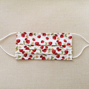 May include: White face mask with red strawberries printed on it. The mask has white elastic straps.