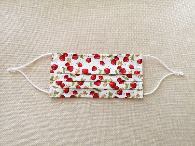 May include: White face mask with red strawberries printed on it. The mask has white elastic straps.