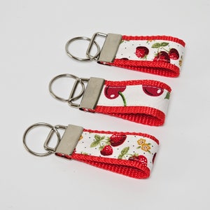 May include: Three keyrings with a red strap and a white fabric overlay featuring a fruit pattern. The fabric has a pattern of cherries and strawberries. Each keyring has a silver metal ring and clasp.