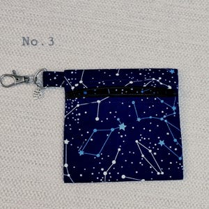 May include: A square fabric pouch in dark blue featuring a white and light blue constellation and star pattern. The pouch has a black zip closure, a silver metal lobster clasp, and a small heart-shaped charm with the text No. 3 visible.