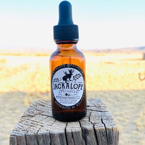May include: A brown glass bottle with a black dropper, labelled "JACKALOPE APOTHECARY." The label features a jackalope illustration. The bottle sits on a weathered wooden post against a blurred outdoor background.
