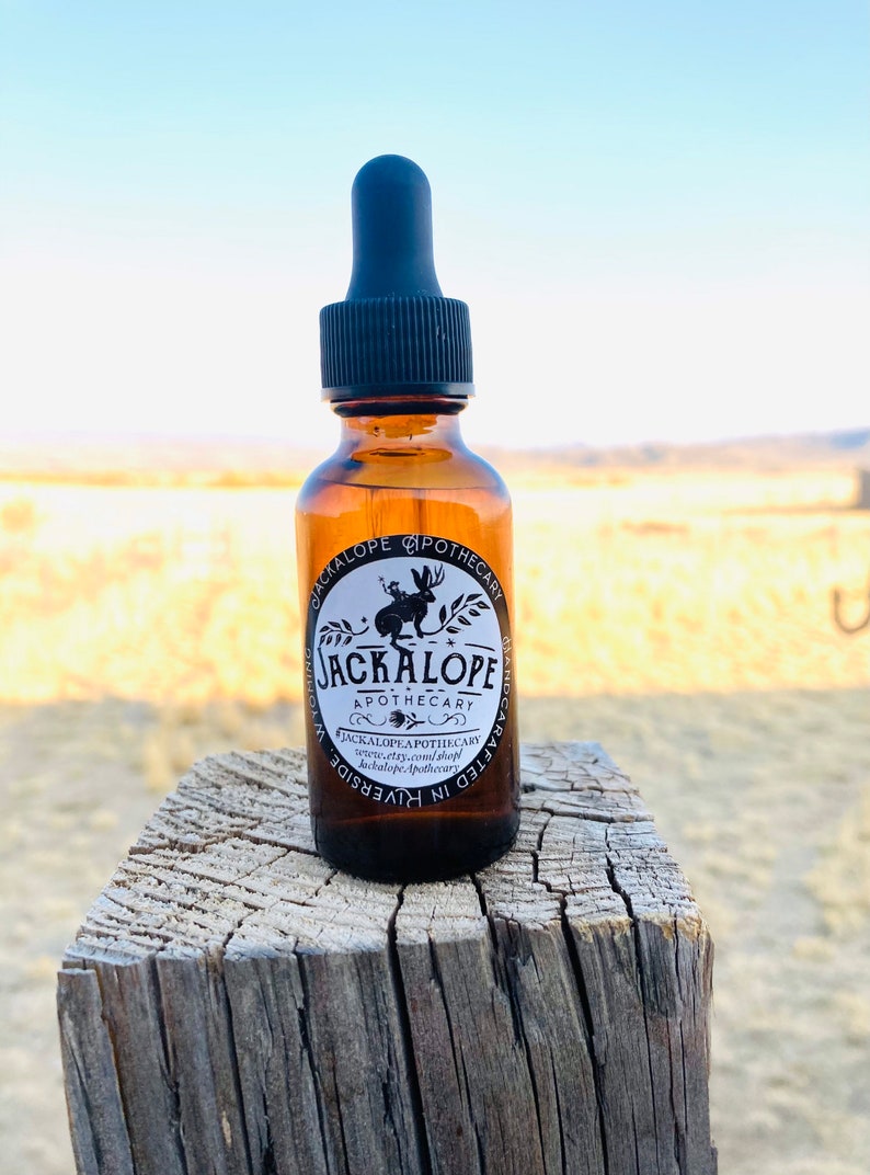 May include: A brown glass bottle with a black dropper, labelled "JACKALOPE APOTHECARY." The label features a jackalope illustration. The bottle sits on a weathered wooden post against a blurred outdoor background.
