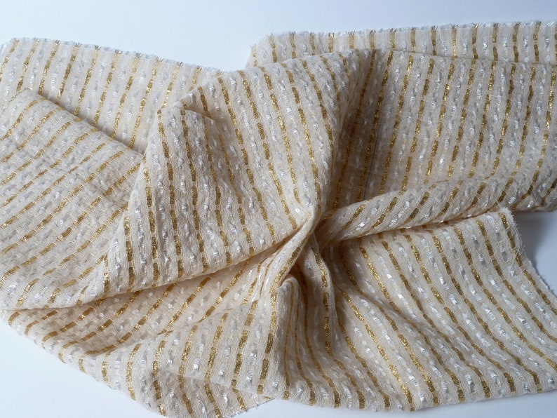 May include: A beige fabric with gold and silver stripes. The fabric has a textured surface and is woven with a subtle shimmer.