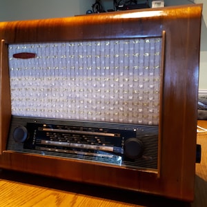 K&ouml;nnte beinhalten: Ein vintage Holzradio mit einem wei&szlig;en Stoff-Lautsprechergitter und schwarzen Drehkn&ouml;pfen. Das Radio hat einen Lautst&auml;rkeregler mit der Aufschrift "Volume" und einen Drehknopf mit der Aufschrift "Tuning".