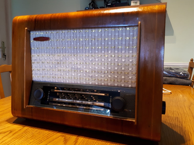K&ouml;nnte beinhalten: Ein vintage Holzradio mit einem wei&szlig;en Stoff-Lautsprechergitter und schwarzen Drehkn&ouml;pfen. Das Radio hat einen Lautst&auml;rkeregler mit der Aufschrift "Volume" und einen Drehknopf mit der Aufschrift "Tuning".