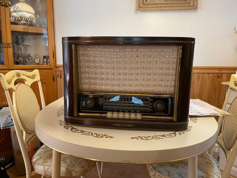 K&ouml;nnte beinhalten: Ein vintage Holzradio mit einem braunen Geh&auml;use und einer goldfarbenen Frontplatte. Das Radio hat ein Lautsprechergitter mit einem wei&szlig;en Netz und ein Zifferblatt mit schwarzem Hintergrund und wei&szlig;en Markierungen. Das Radio steht auf einem wei&szlig;en Tisch mit einem dekorativen Muster.