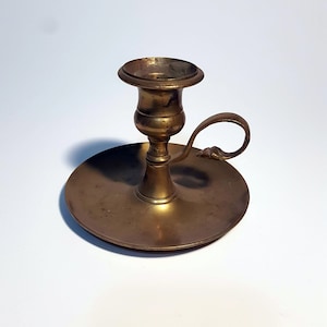 May include: A brass candlestick with a round base and a single candle holder. The candlestick has a small handle on the side.
