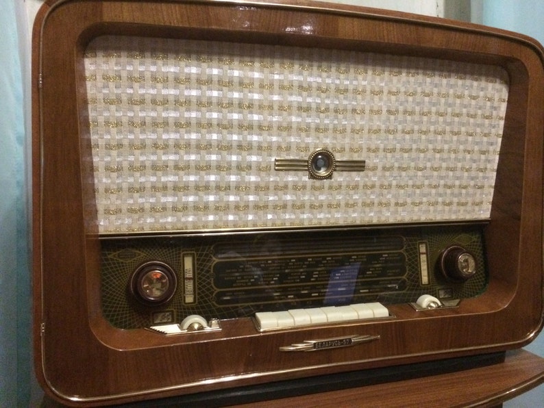 May include: A vintage wooden radio with a white and gold speaker grille, a glass dial with a black and gold design, and a row of white buttons. The radio has a brown wooden cabinet with a gold trim.