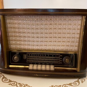 K&ouml;nnte beinhalten: Ein vintage Holzradio mit braunem Geh&auml;use und goldener Verzierung. Das Radio hat ein Lautsprechergitter mit Stoffbezug und ein Zifferblatt mit Zahlen und Buchstaben. Das Radio hat zwei Kn&ouml;pfe an der Vorderseite und ein Typenschild an der Unterseite.