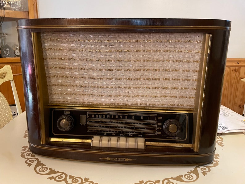 K&ouml;nnte beinhalten: Ein vintage Holzradio mit braunem Geh&auml;use und goldener Verzierung. Das Radio hat ein Lautsprechergitter mit Stoffbezug und ein Zifferblatt mit Zahlen und Buchstaben. Das Radio hat zwei Kn&ouml;pfe an der Vorderseite und ein Typenschild an der Unterseite.