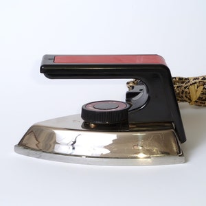 May include: A vintage black and silver iron with a red handle and a round temperature control knob. The iron has a cord with a braided fabric covering.