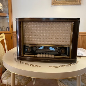 May include: A vintage wooden radio with a brown cabinet and a gold-coloured front panel. The radio has a speaker grille with a white mesh and a dial with a black background and white markings. The radio is sitting on a white table with a decorative pattern.