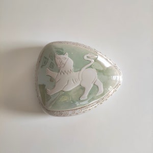 May include: A ceramic trinket box with a green glaze and a white lion design. The box is heart-shaped and has a decorative border.