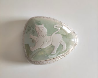 Könnte beinhalten: Eine Keramik-Schmuckschatulle mit grüner Glasur und einem weißen Löwenmotiv. Die Schachtel ist herzförmig und hat einen dekorativen Rand.