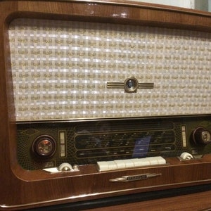 K&ouml;nnte beinhalten: Ein Vintage-Holzradio mit einem wei&szlig;en und goldenen Lautsprechergitter, einem Glaszifferblatt mit schwarzem und goldenem Design und einer Reihe wei&szlig;er Kn&ouml;pfe. Das Radio hat ein braunes Holzkabinett mit goldener Verzierung.