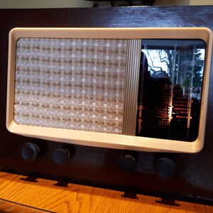 K&ouml;nnte beinhalten: Ein vintage Holzradio mit wei&szlig;em Zifferblatt und schwarzen Kn&ouml;pfen. Das Radio hat ein Lautsprechergitter und ein Glasfenster, das die inneren Mechanismen des Radios zeigt.