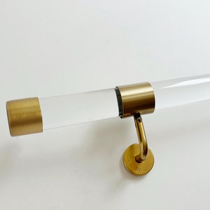 Op de afbeelding: Een transparante acryl handrail met gouden metalen eindkappen en een gouden metalen wandbevestiging. De wandbevestiging heeft het nummer "191" gegraveerd.