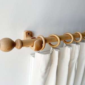 May include: A wooden curtain pole with finials and rings. The pole is mounted to a white wall and has white curtains hanging from it.
