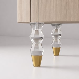 May include: A light wood cabinet base featuring two clear acrylic furniture legs. Each leg has a faceted, stacked geometric design with a metallic gold conical tip at the bottom and a visible silver threaded bolt connecting to the cabinet frame.