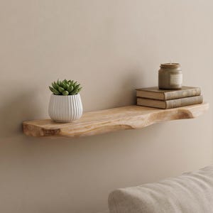Peut inclure: Une étagère flottante en bois à bord naturel fixée au mur. L'étagère présente un grain de bois brun clair avec un bord avant irrégulier et organique. Un pot en céramique blanc nervuré avec une plante succulente verte et deux livres empilés.