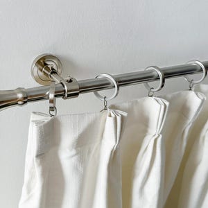 May include: A brushed nickel curtain rail with white curtains. The rail has decorative end pieces and is mounted on a white wall. The curtains are hanging from metal rings.