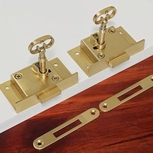 May include: Two brass keyhole escutcheons with a key in each. The escutcheons are mounted on a white surface and a wooden surface.